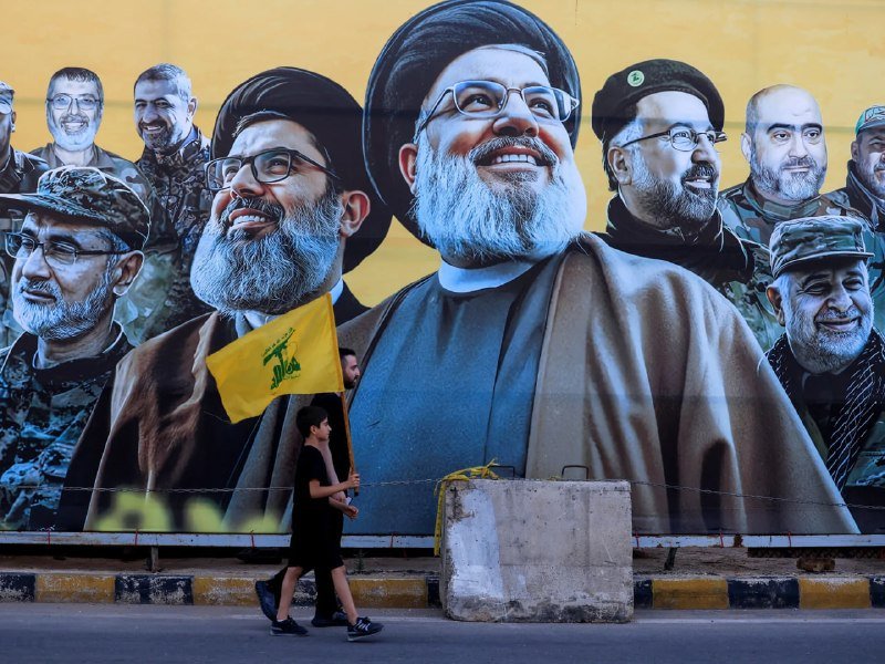 A woman carrying a yellow flag walks past a large outdoor mural depicting several bearded men in black turbans and military attire.