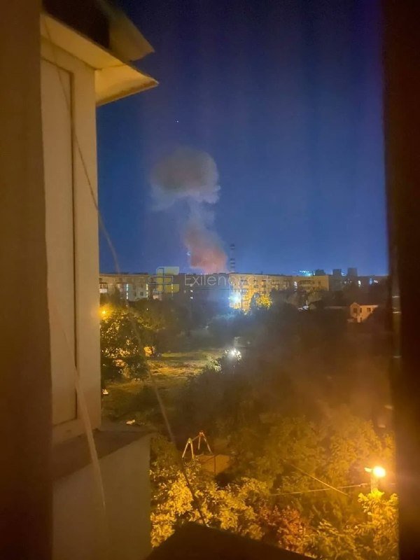A nighttime cityscape viewed through a window shows a large plume of smoke rising from a bright fire among residential buildings.