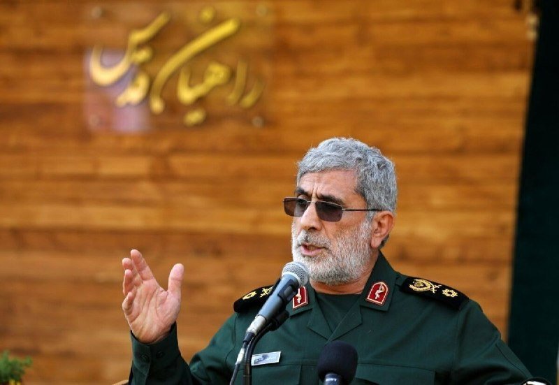 A gray-bearded man in a green military uniform with insignia speaks into a microphone, gesturing with one hand, against a wooden wall with Arabic calligraphy.