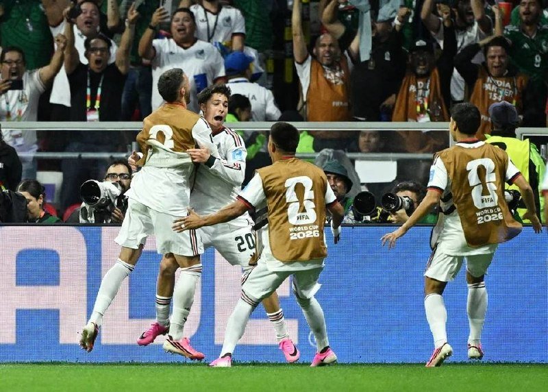 Soccer players in white jerseys and gold "FIFA World Cup 2026" bibs celebrate a goal, while cheering fans and photographers are visible in the background.