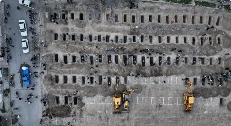 Aerial view of a dirt lot with rows of rectangular excavated graves, workers, and two yellow excavators digging additional plots.