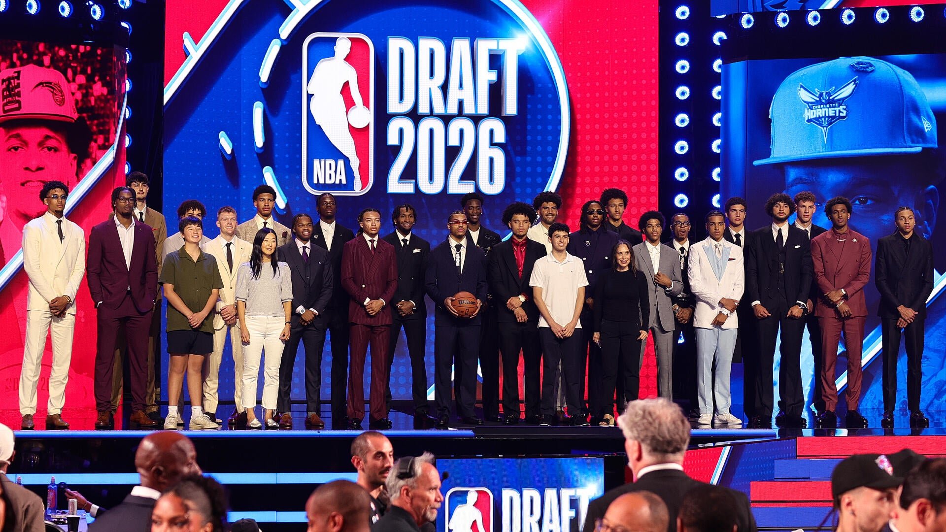 A large group of formally dressed young men and women pose together on stage in front of a brightly lit "NBA Draft 2026" logo, with a basketball held at center.