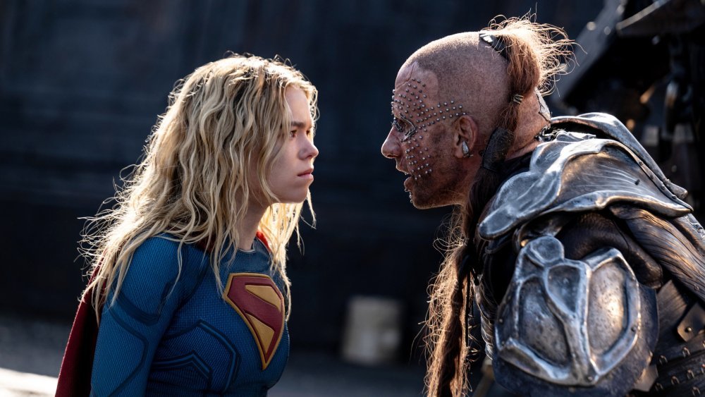 A blonde woman in a blue and red suit with an "S" emblem faces a metal-studded, armored man with a partially shaved head in a dimly lit industrial setting.