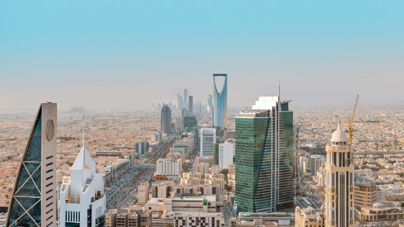 Aerial view of a dense Middle Eastern city skyline featuring modern skyscrapers, including a distinctive tower with a large rectangular opening near its top, under a hazy sky.