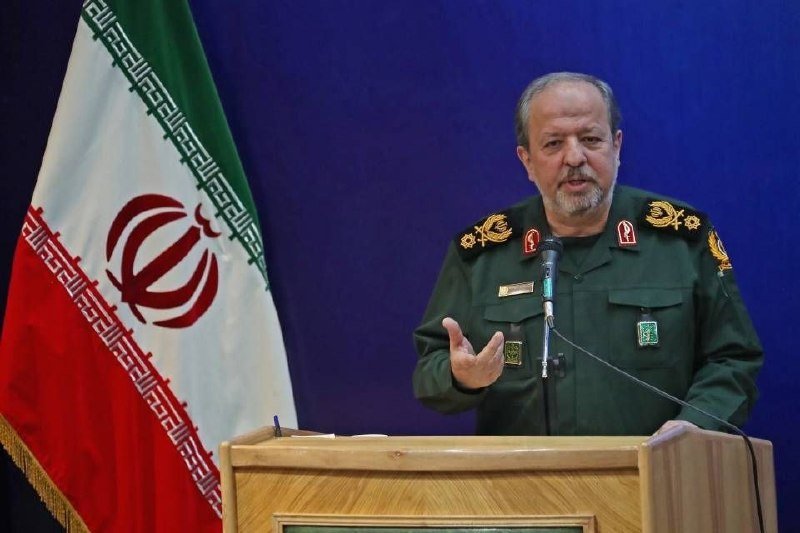 A man in a military uniform gestures with his hand while speaking at a wooden podium, with an Iranian flag visible to his left against a dark blue backdrop.