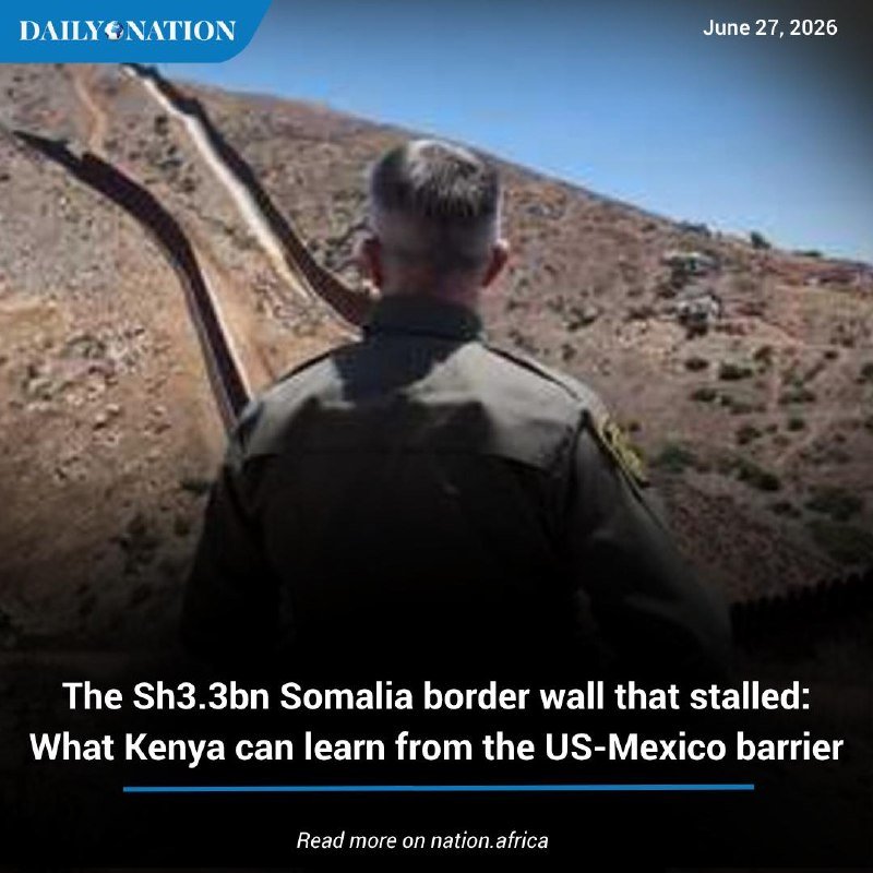 A uniformed agent viewed from behind looks out over an arid landscape toward a tall border barrier wall stretching across the terrain under a clear blue sky.