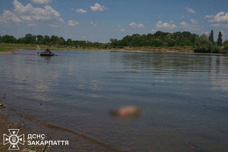 A person rows a small inflatable boat on a wide river under a partly cloudy sky, with a blurred figure visible in the water near the sandy shore.