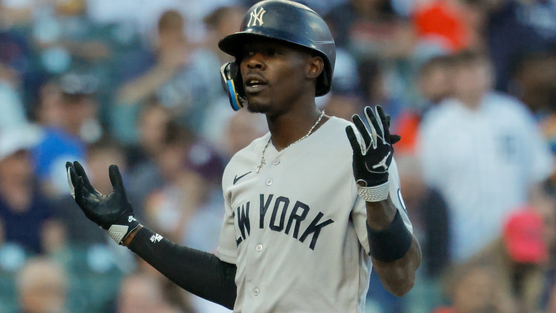 A baseball player in a white New York uniform and navy batting helmet gestures with both hands raised, wearing black batting gloves during a game.