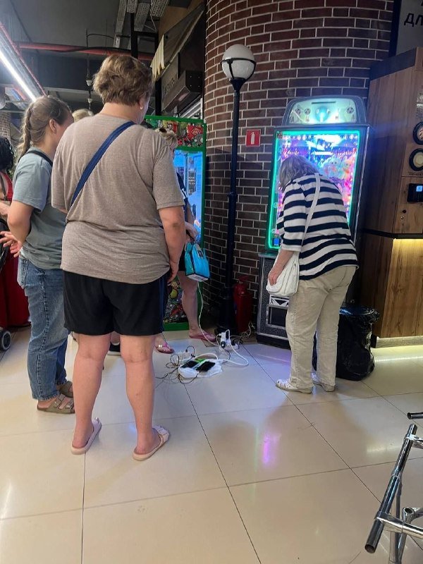 Several people stand near a glowing arcade game machine with a brick wall and tiled floor in the background, with cables and a power strip visible on the ground.