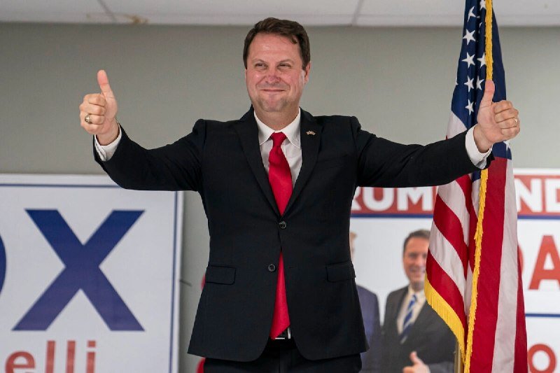 A man in a dark suit and red tie gives two thumbs up while standing in front of an American flag and campaign banners displaying partial text and another person's image.