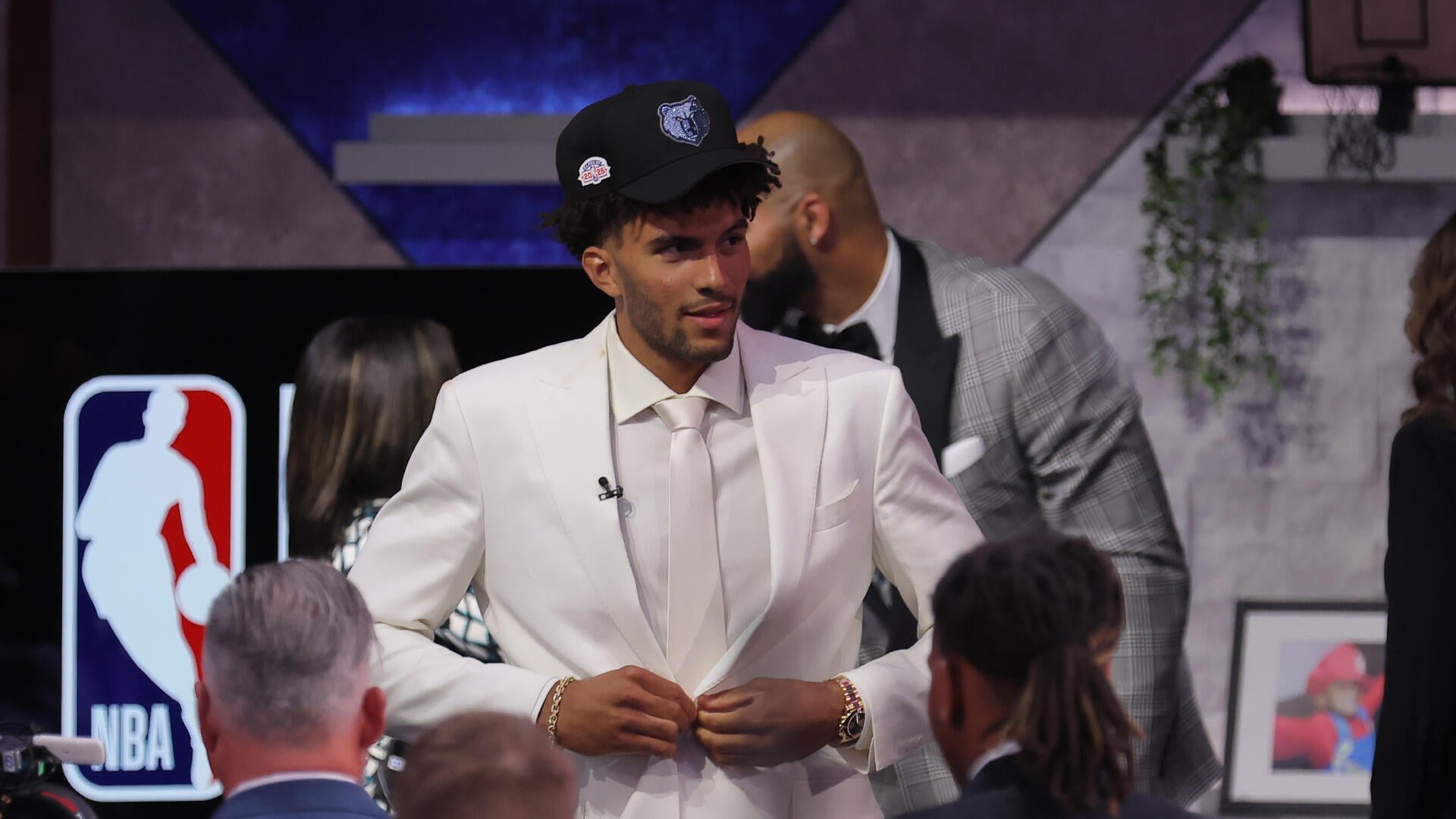 A young man in a white suit and a black Memphis Grizzlies cap buttons his jacket on stage beside an NBA logo, surrounded by other formally dressed attendees.