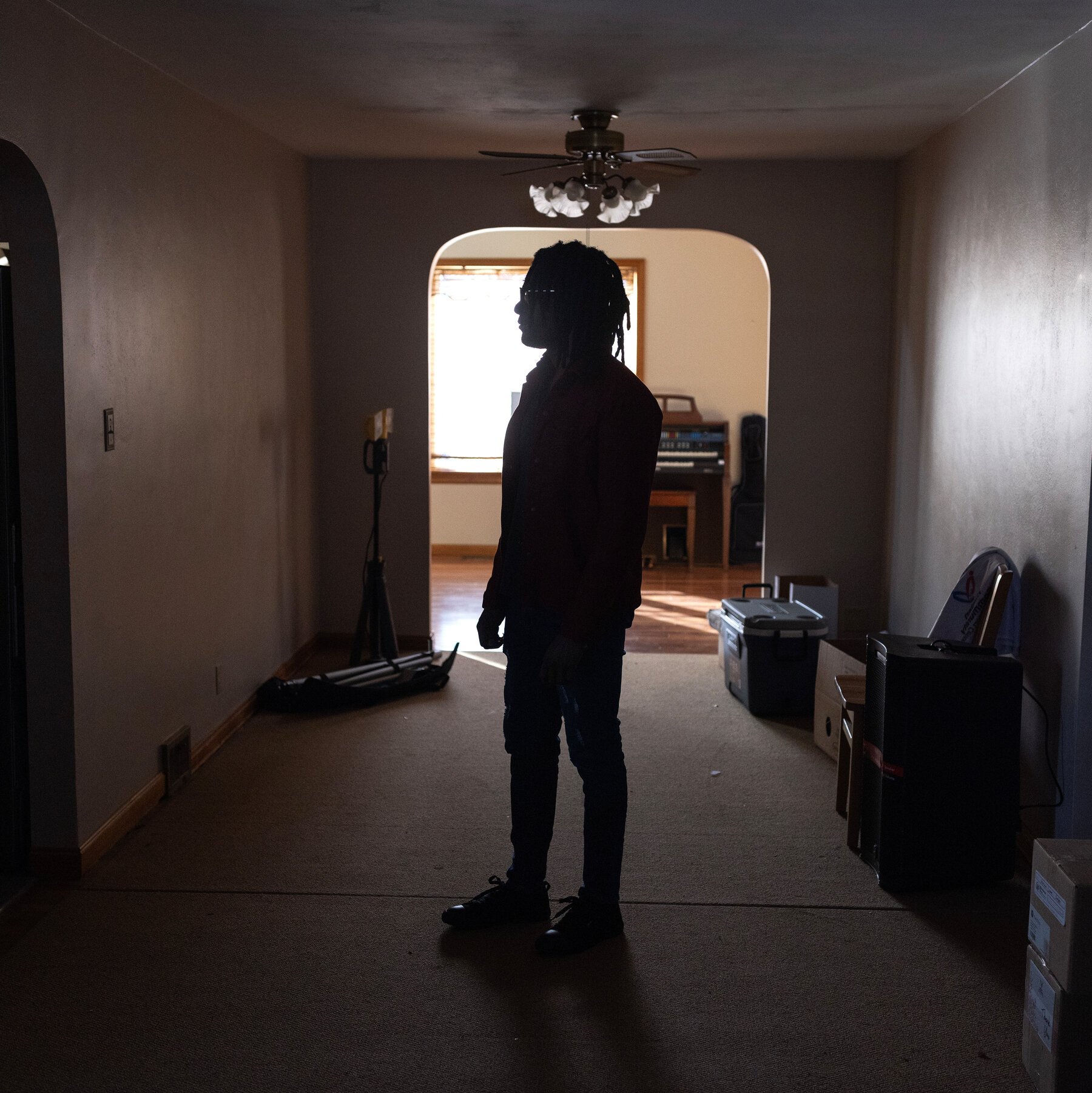 A silhouetted person with dreadlocks stands in a dimly lit hallway facing a backlit room, surrounded by cardboard boxes and storage containers.