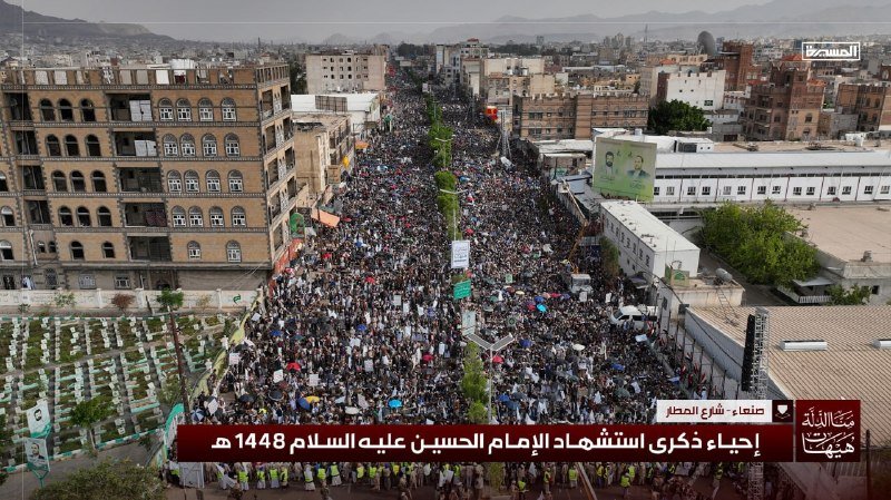 A massive crowd fills a wide urban street lined with multi-story buildings during a daytime commemoration event, with Arabic text overlays visible.