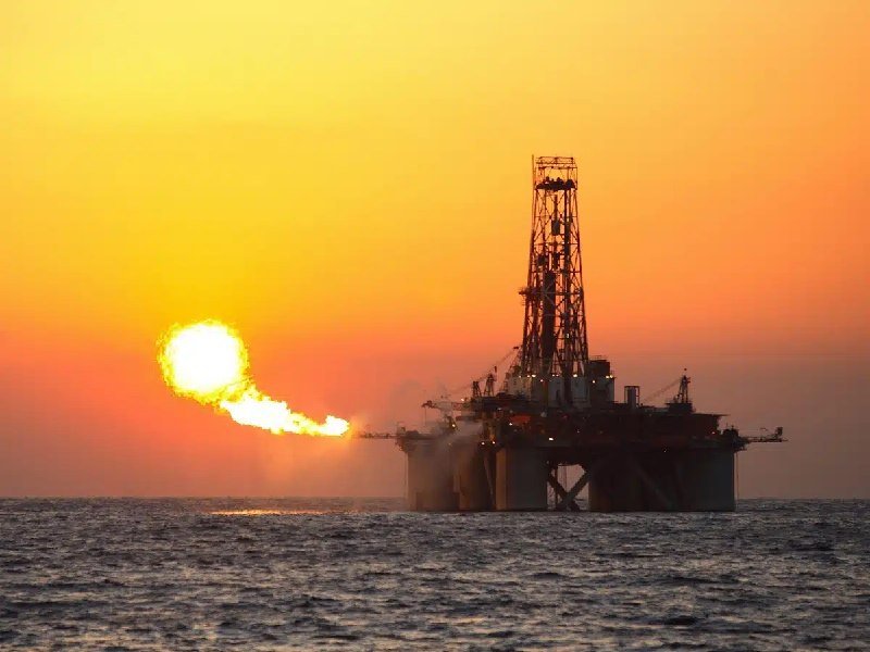 An offshore oil drilling rig sits on the ocean at sunset while a gas flare burns brightly from its side.
