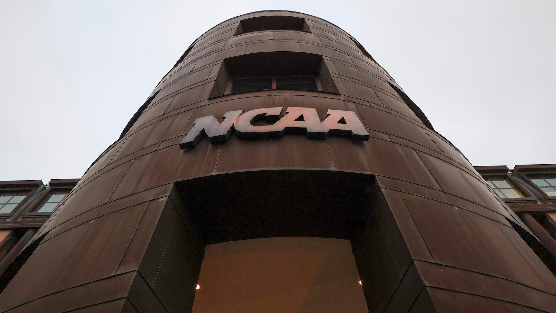 A low-angle view shows a dark brown, rocket-shaped architectural structure displaying the raised "NCAA" logo against a pale sky.