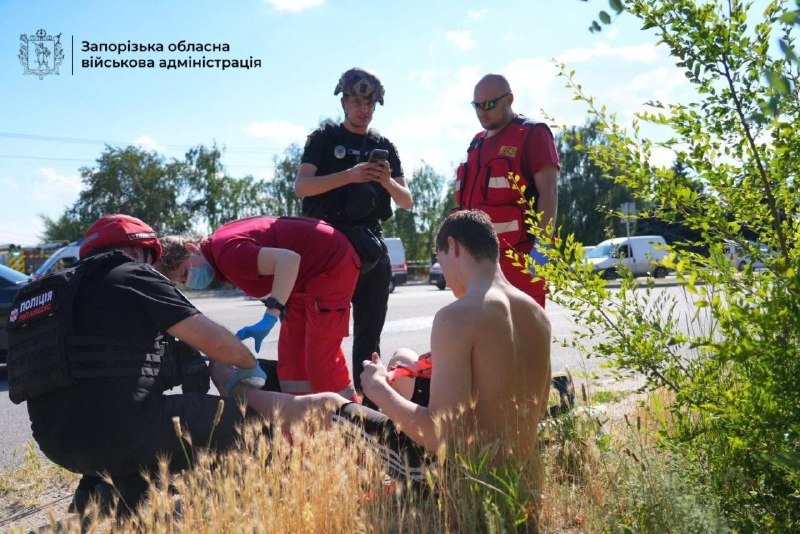 Emergency responders in red uniforms and black tactical gear attend to a shirtless man lying on the ground beside a road, with parked vehicles visible in the background.
