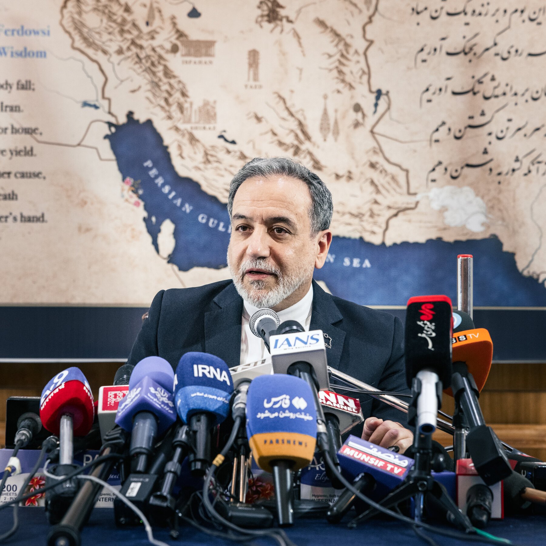 A gray-bearded man in a dark suit speaks at a press conference behind a cluster of labeled media microphones, with a map showing the Persian Gulf and Arabian Sea on the wall behind him.