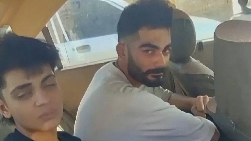 A bearded man in a white shirt sits in the driver's seat of a car beside a younger passenger.