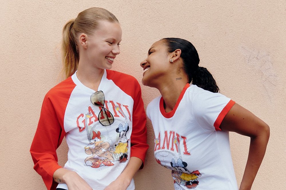 Two women in white and red Disney-themed "Ganni" shirts smile and look at each other against a beige textured wall.