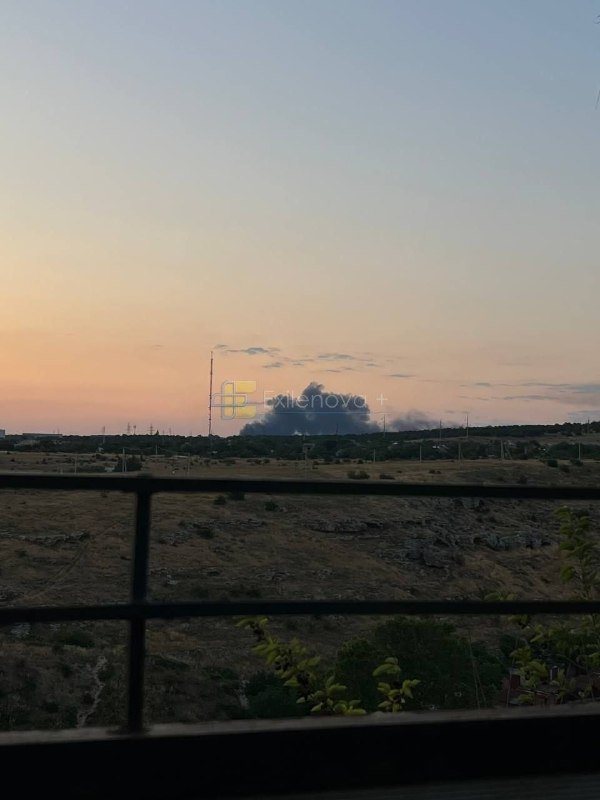 A sunset sky over a dry, brush-covered landscape with distant smoke rising beyond a foreground railing.