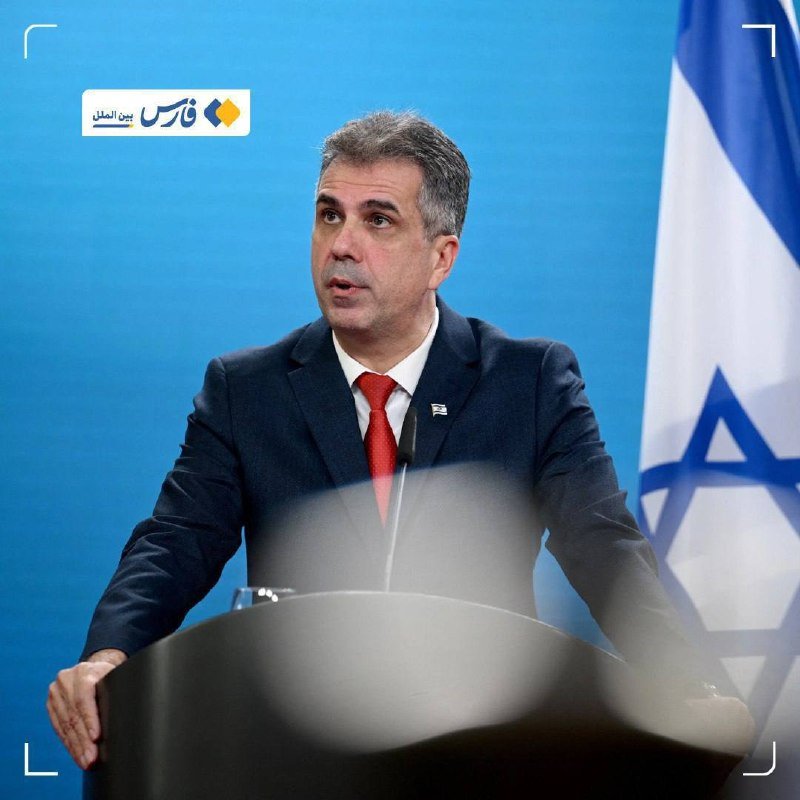 A man in a dark suit and red tie speaks at a podium with microphones, standing before a blue backdrop and an Israeli flag.