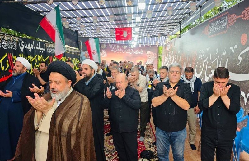 Men in clerical and civilian attire stand in prayer with raised hands beneath a striped tent, flanked by Iranian flags and Persian/Arabic banners.