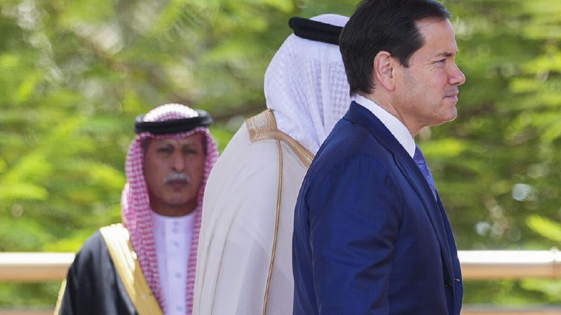 Three men in formal attire—two in traditional Arab headdress and robes, one in a dark suit—stand outdoors against a blurred green background.