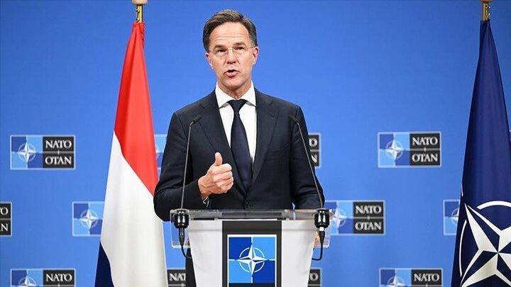 A man in a dark suit speaks at a podium bearing the NATO emblem, gesturing with one hand, flanked by a red-white-blue flag and a blue NATO flag.