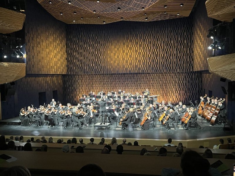 A full orchestra in formal attire performs on a wood-paneled concert hall stage, conducted by a standing figure, with audience members visible in the foreground.