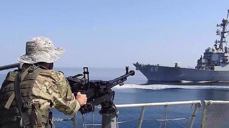 A soldier in camouflage and boonie hat stands behind a mounted machine gun on a ship, aiming toward a gray naval destroyer marked "68" on the horizon.