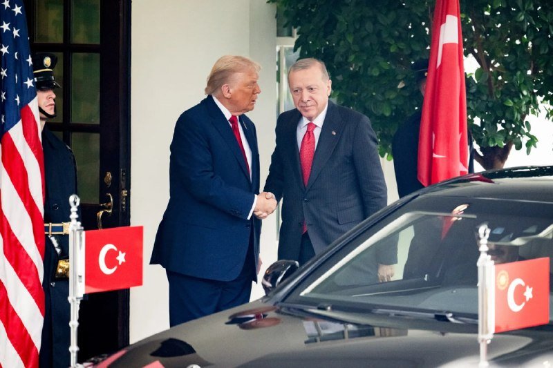 Two men in suits shake hands beside a black car, flanked by U.S. and Turkish flags.