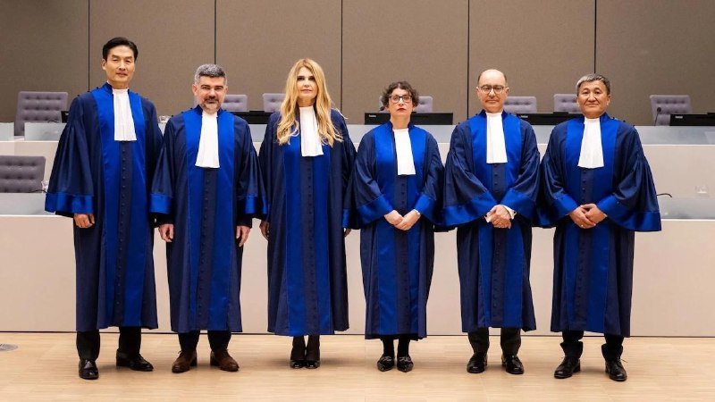 Seven people in blue judicial robes with white collars stand in a line inside a formal chamber with empty chairs behind them.