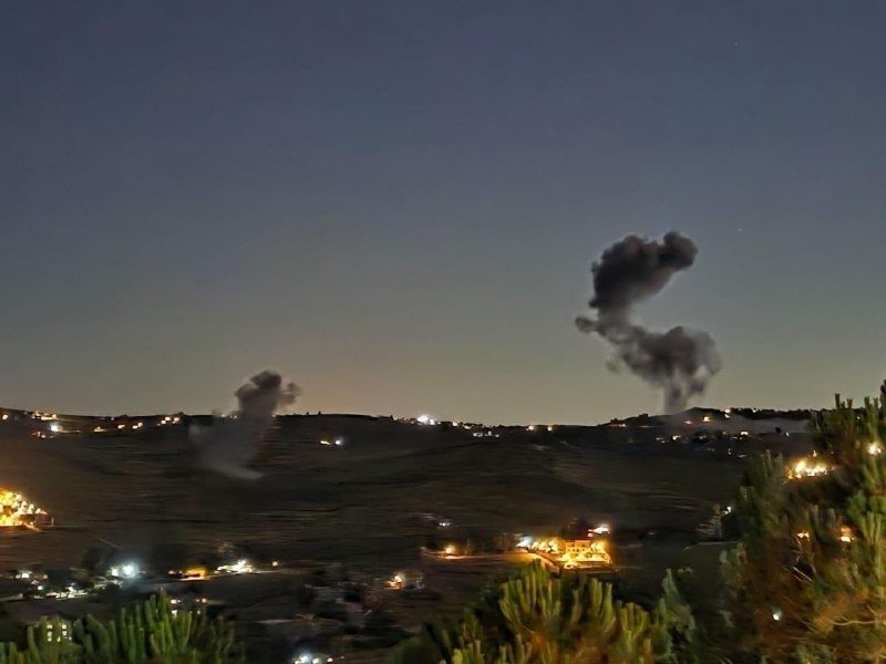 Two plumes of dark smoke rise from a hilly landscape dotted with scattered lights under a twilight sky.