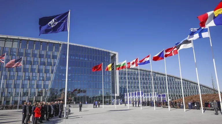 A NATO flag flies alongside multiple national flags in front of a large glass office building, with formally dressed people gathered in the plaza below.