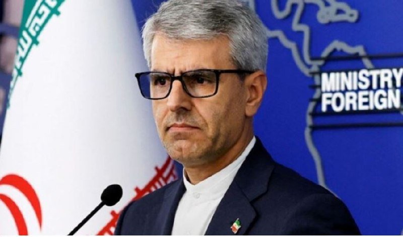 A gray-haired man in glasses and a dark suit stands at a microphone in front of an Iranian flag and a blue backdrop labeled "MINISTRY OF FOREIGN."