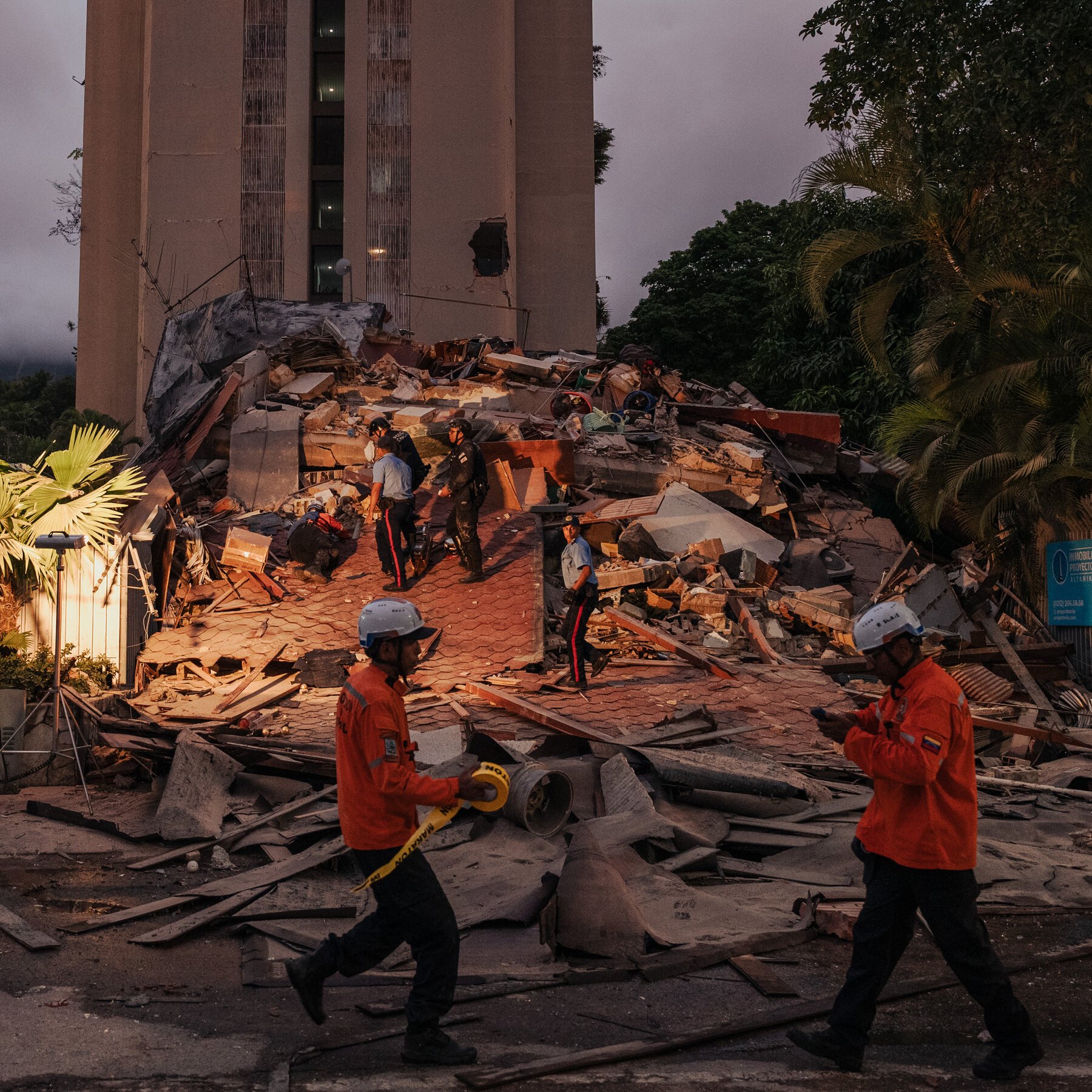 Rescue workers in orange uniforms and helmets walk through rubble near a partially collapsed building at dusk, with palm trees in the background.