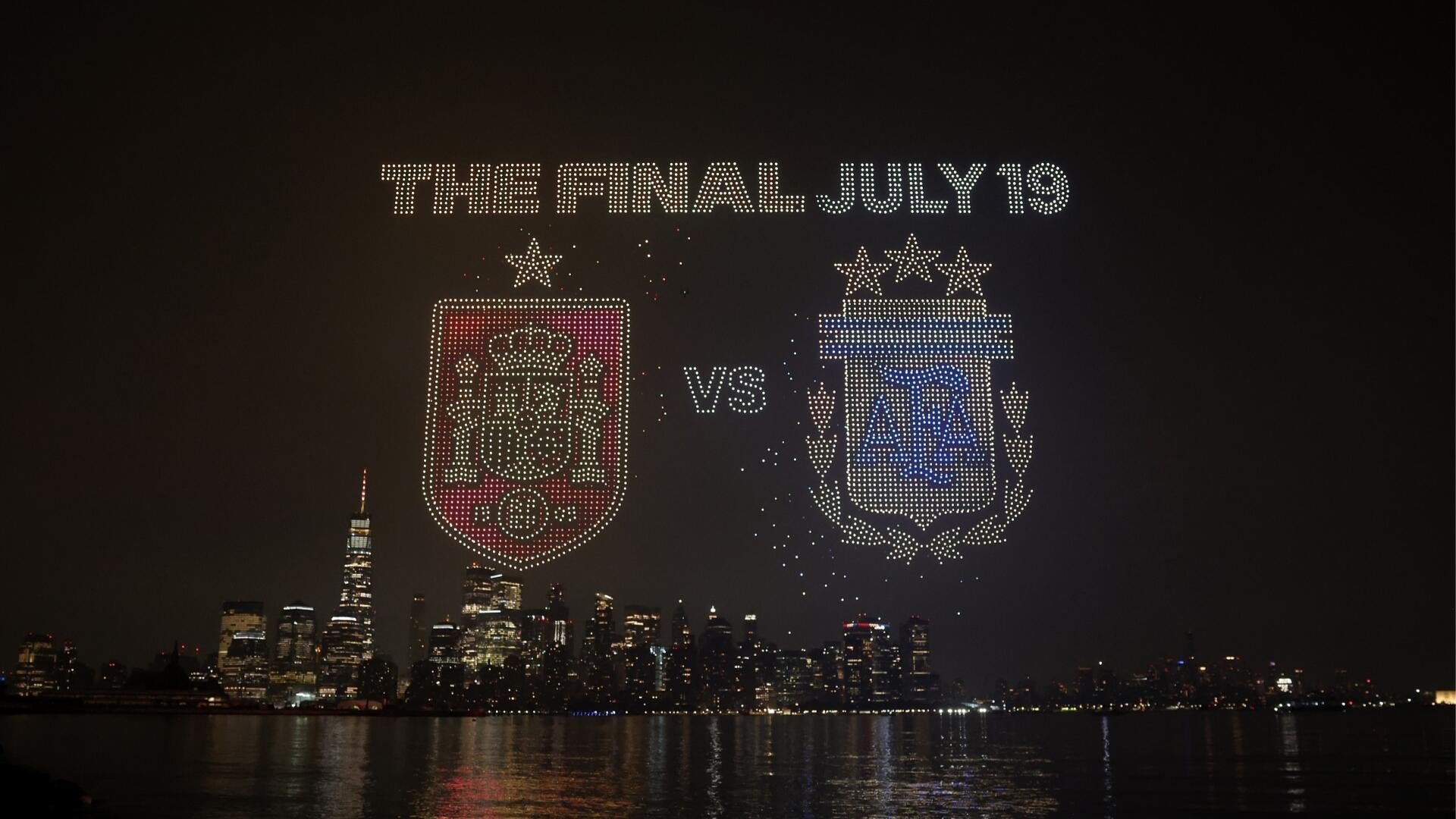 A drone light show displays "THE FINAL - JULY 19" above two team crests separated by "VS" over a nighttime city skyline and waterway.