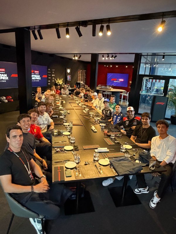 A large group of people sits along a long dining table set with plates and glasses inside a modern venue, with screens displaying "BELGIAN GRAND PRIX 2025" visible on the walls.