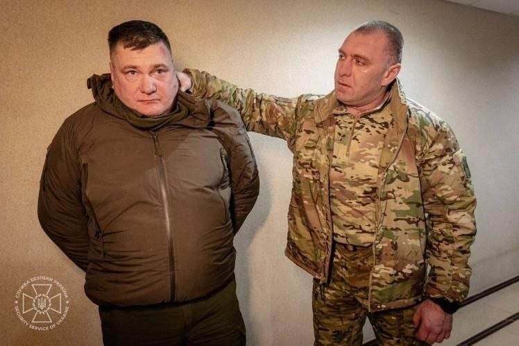 Two men stand against a plain wall; one in a dark puffy jacket faces forward with hands behind his back, while the other in camouflage military attire holds him from behind.