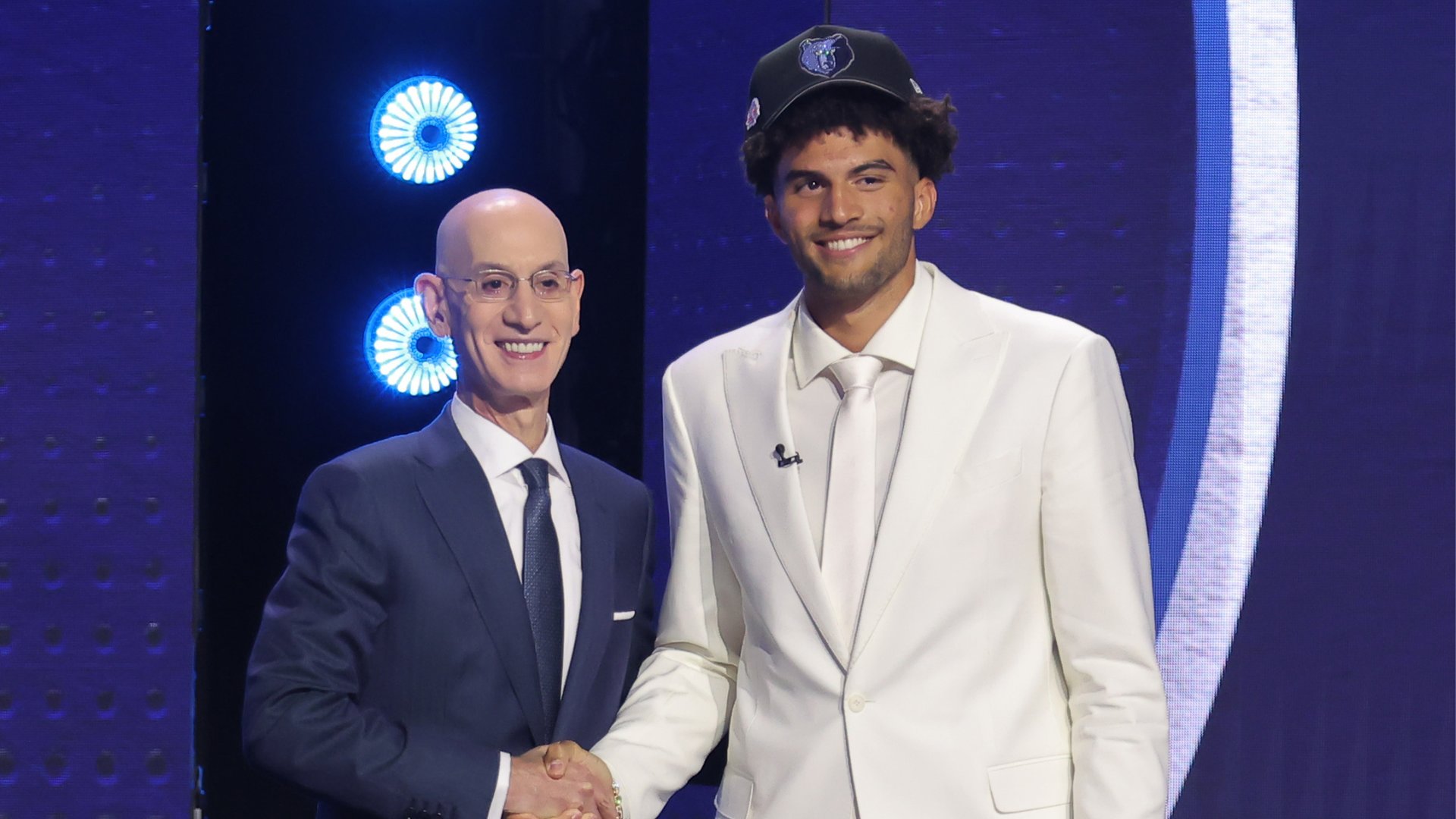A bald man in a dark suit shakes hands with a taller young man wearing a white suit and a dark baseball cap on a blue-lit stage.