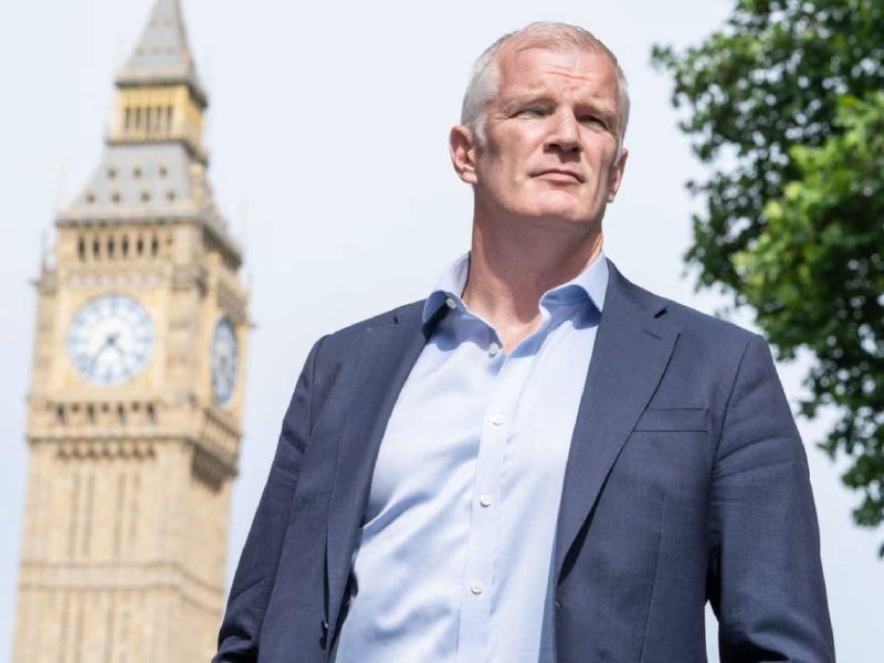 A gray-haired man in a blue blazer and light blue shirt stands outdoors, with the iconic Big Ben clock tower visible in the background.
