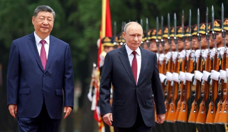 Two men in dark suits and ties walk past a row of uniformed honor guards standing at attention with rifles.