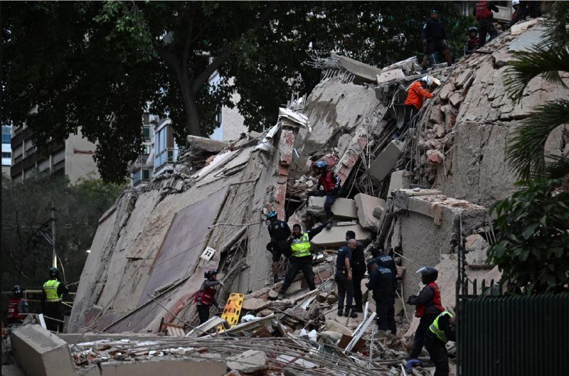 Rescue workers in helmets and high-visibility vests climb over a large pile of concrete and building debris from a collapsed structure.