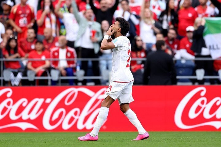 A soccer player in a white uniform with pink cleats stands on a grass field, blowing a kiss toward cheering fans in red behind a Coca-Cola advertising board.