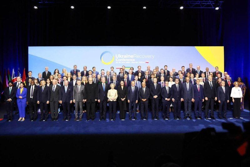 Dozens of formally dressed delegates pose on stage in front of a backdrop reading "Ukraine Recovery Conference" with dates and flags visible.