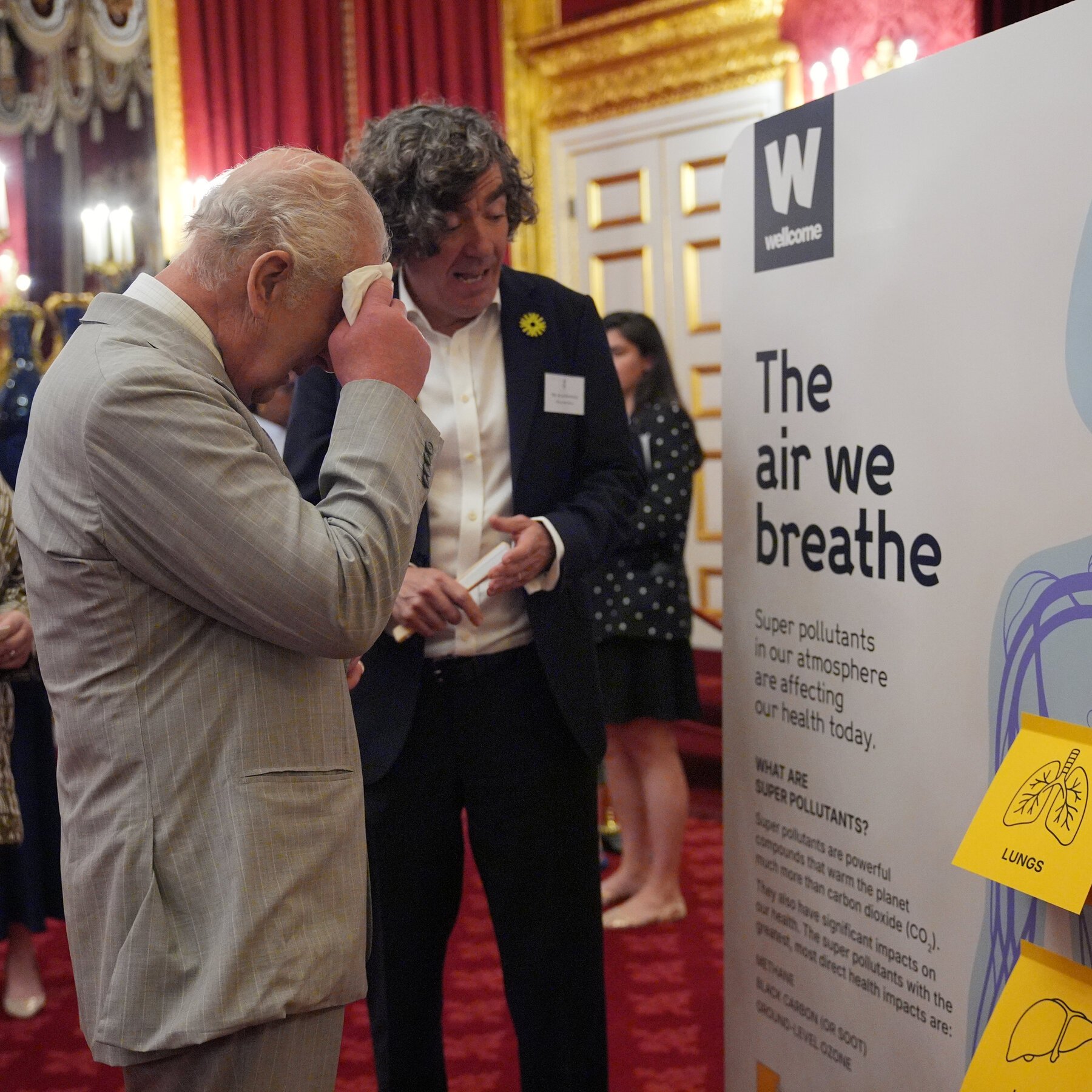 Two men in suits stand near a "Wellcome" exhibition sign reading "The air we breathe," with one man wiping his face with a handkerchief.