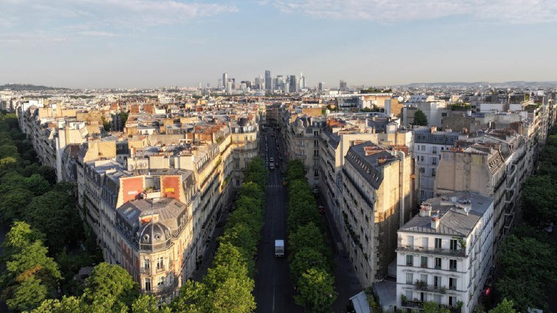 Aerial view of a tree-lined city avenue flanked by multi-story residential buildings, with a cluster of modern skyscrapers visible on the distant horizon under a partly cloudy sky.