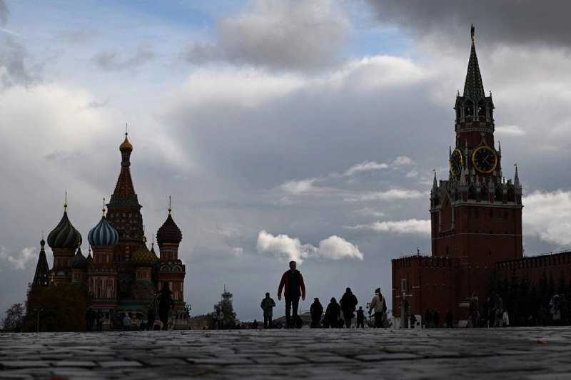Silhouetted people walk across a cobblestone square between an ornate cathedral with colorful onion domes and a tall red clock tower under a cloudy sky.