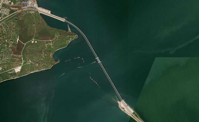 Satellite image showing a long bridge crossing a body of dark water, connecting a developed coastal landmass on the left to a smaller land area on the lower right.