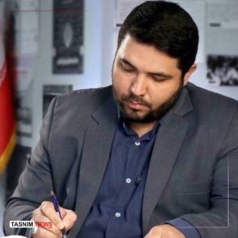 A bearded man in a gray suit and blue shirt sits writing with a pen, looking downward, with a "Tasnim News" watermark visible.